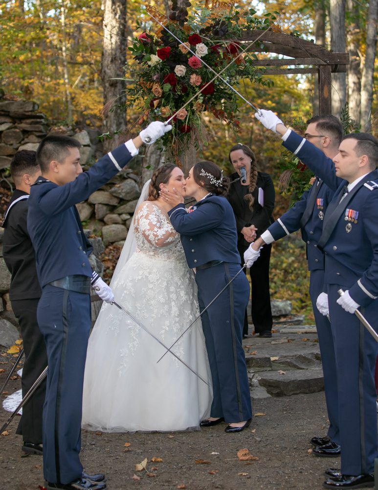 A bride and groom kiss as two men in uniform hold swords.